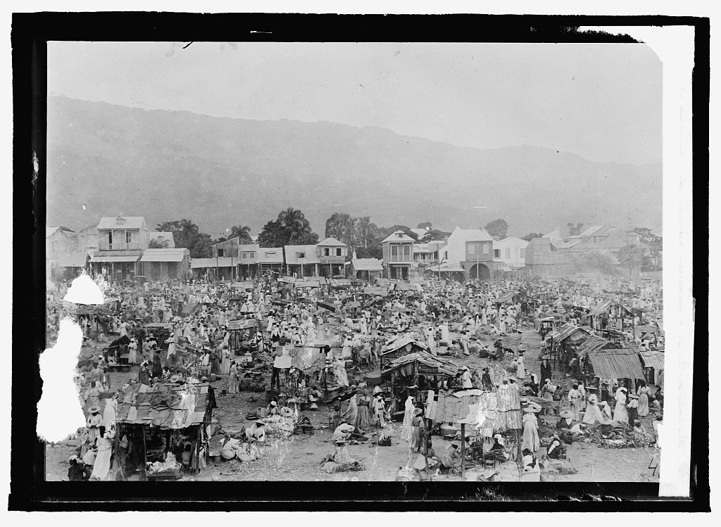 Historical black-and-white view of a crowded market with stalls, low buildings and hills beyond; the glass-negative image has visible damage.
