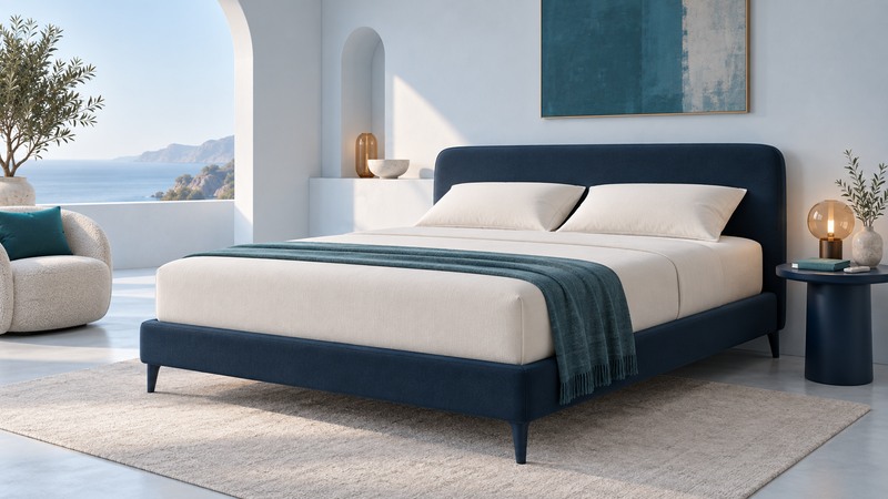 An unbranded ivory bed with a navy frame stands beside a remote and teal notebook on a bedside table.